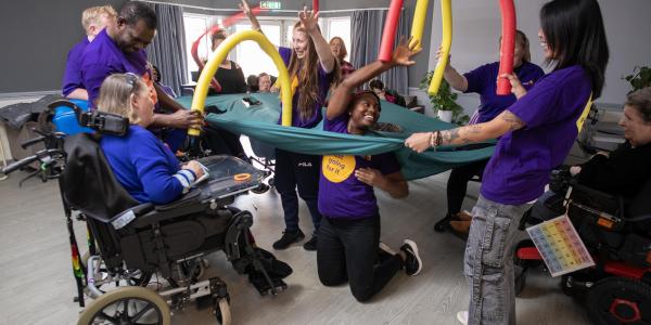 A group of disabled residents in a living area playing with the care workers with foam tubing who are wearing branded t shirts