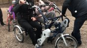A man and a woman sitting in an adapted tricycle with a volunteer standing in front of them