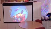 A projector with fish projections with a brown table and chairs in front of it and red, blue, yellow and green foam floor tiles