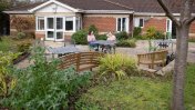 Patio area with shrubs and a wooden bridge and bench with metal framed garden table and chairs with two caucasian females one blond and one brunette sat