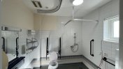 A white tiled bathroom with assistive fittings by the toilet and open shower space