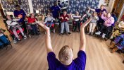 A group of residents in wheelchairs in a circle in a room with oak laminate flooring in front of a caucasian male support worker with blond hair sitting down in front of them with his arms up in the air