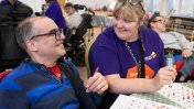 A caucasian male resident with glasses and a blue striped rugby shirt smiling at a caucasian female care worker wearing a purple t shirt both holding dabbers at a bingo game
