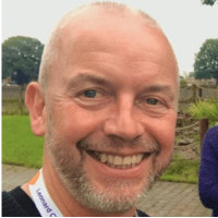 Dave a caucasian male with very short grey hair and stubble smiling at the camera 