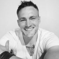 A black and white portrait of Nathan a caucasian male with short dark hair wearing a white t shirt and smiling at the camera with his arms folded