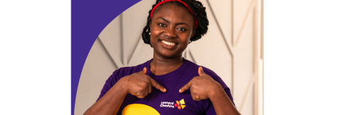 A dark skinned female wearing a branded Leonard Cheshire T shirt and pointing to the logo on it whilst smiling at the camera 