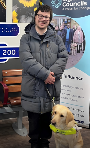 Alex, a white male, has dark hair and glasses and is wearing a long grey duffel coat. Rosie, a golden retriever cross, is by his side.