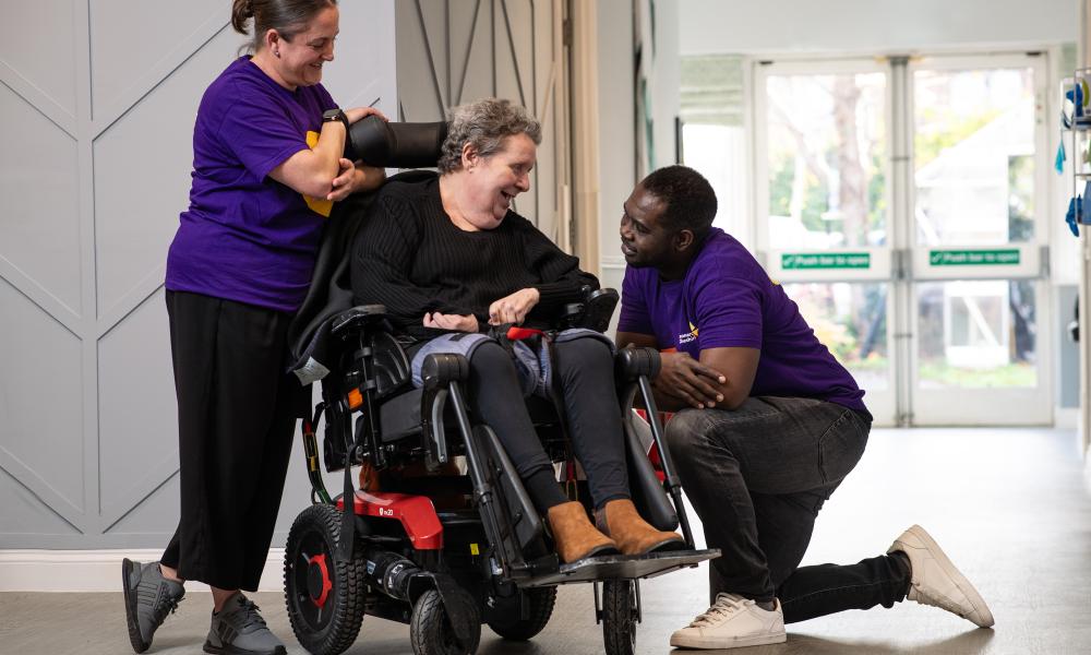A Caucasian female resident in a wheelchair with an african male care worker kneeling beside her on one side and a Caucasian femaie stood next to her on the other side 